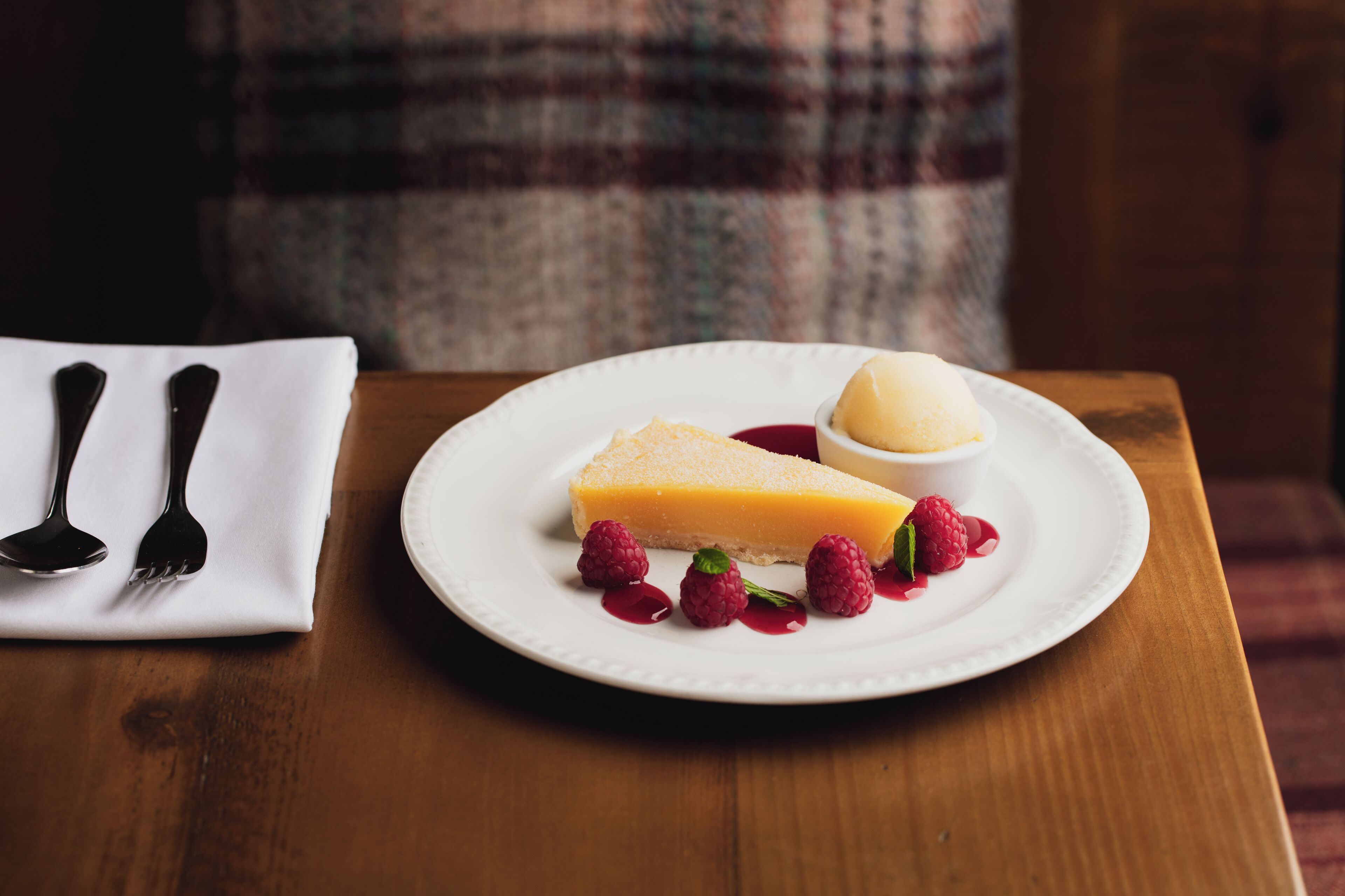 A dessert plate with a slice of cheesecake and raspberries on a white plate, accompanied by a small bowl of ice cream and a napkin with cutlery.