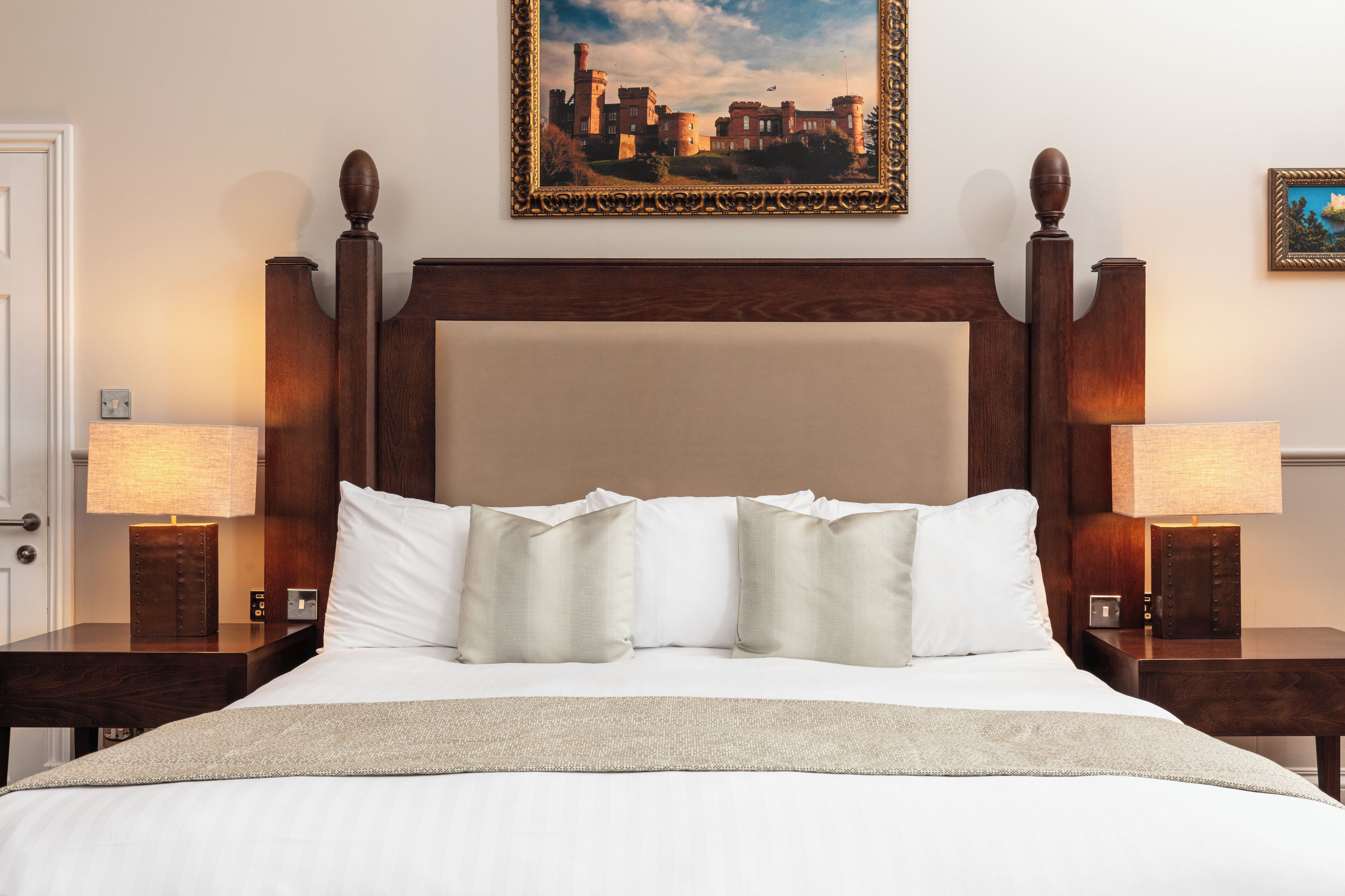 Una cama bien hecha con ropa blanca y un edredón color beige, flanqueada por dos mesitas de noche con lámparas, en una habitación con cabecera de madera y un cuadro en la pared.
