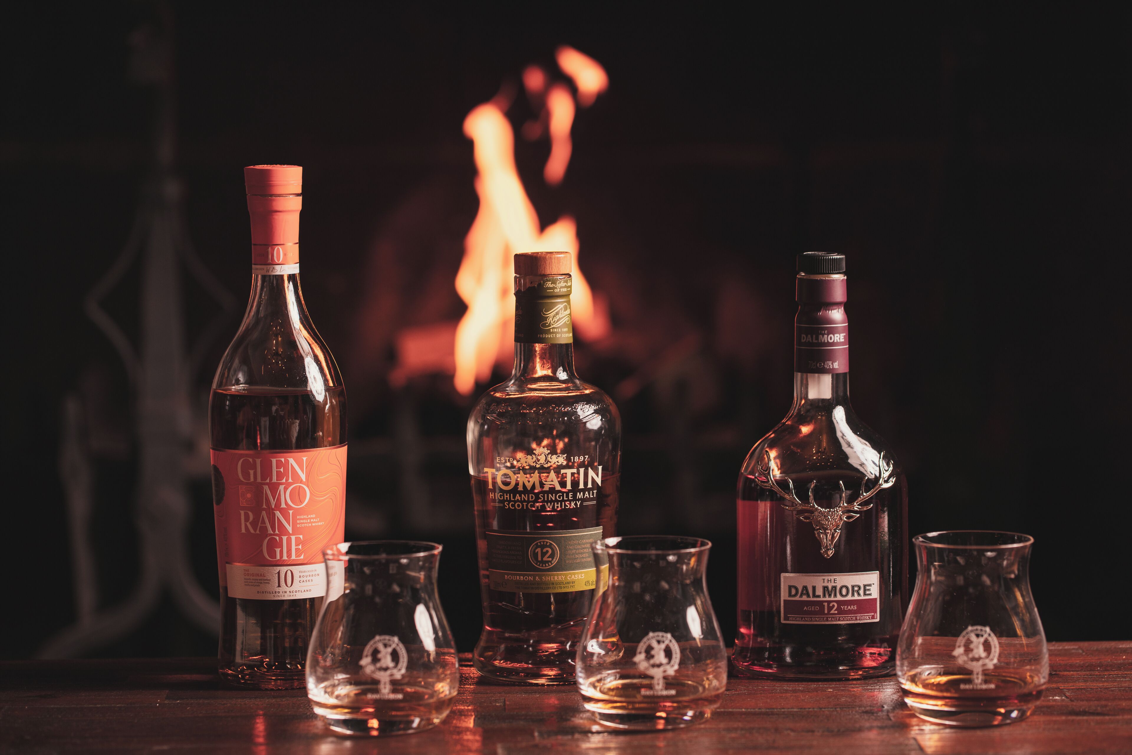 Three bottles of whisky and four glasses on a wooden table with a fireplace in the background.