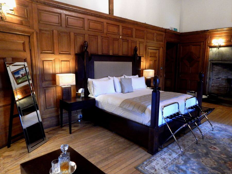 Un espacioso dormitorio con paneles de madera oscura, una cama grande, dos mesitas de noche, un revistero, un espejo, una chimenea y una alfombra estampada.