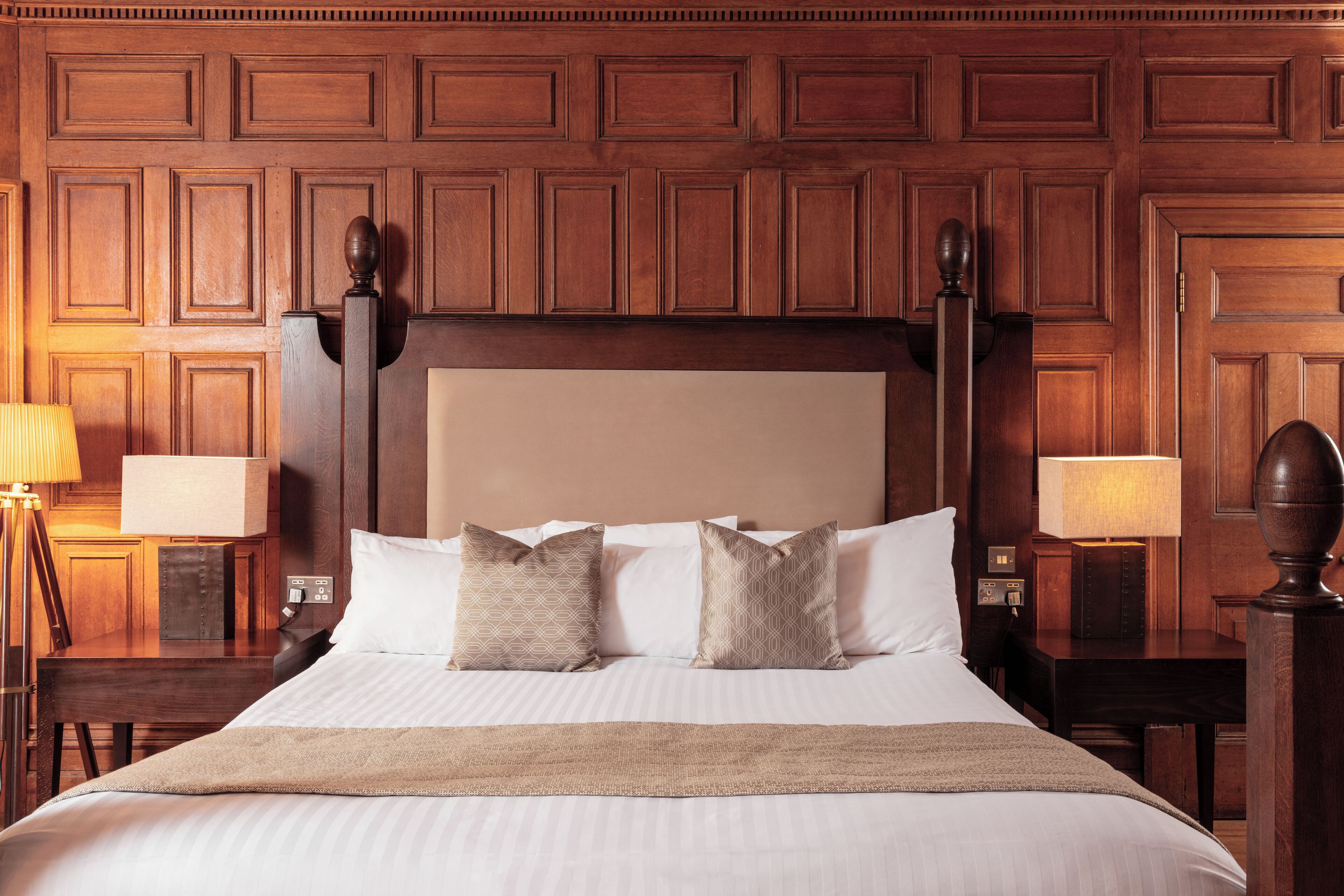 A hotel room with a large bed, wooden headboard, two bedside lamps, and a wooden panel wall.