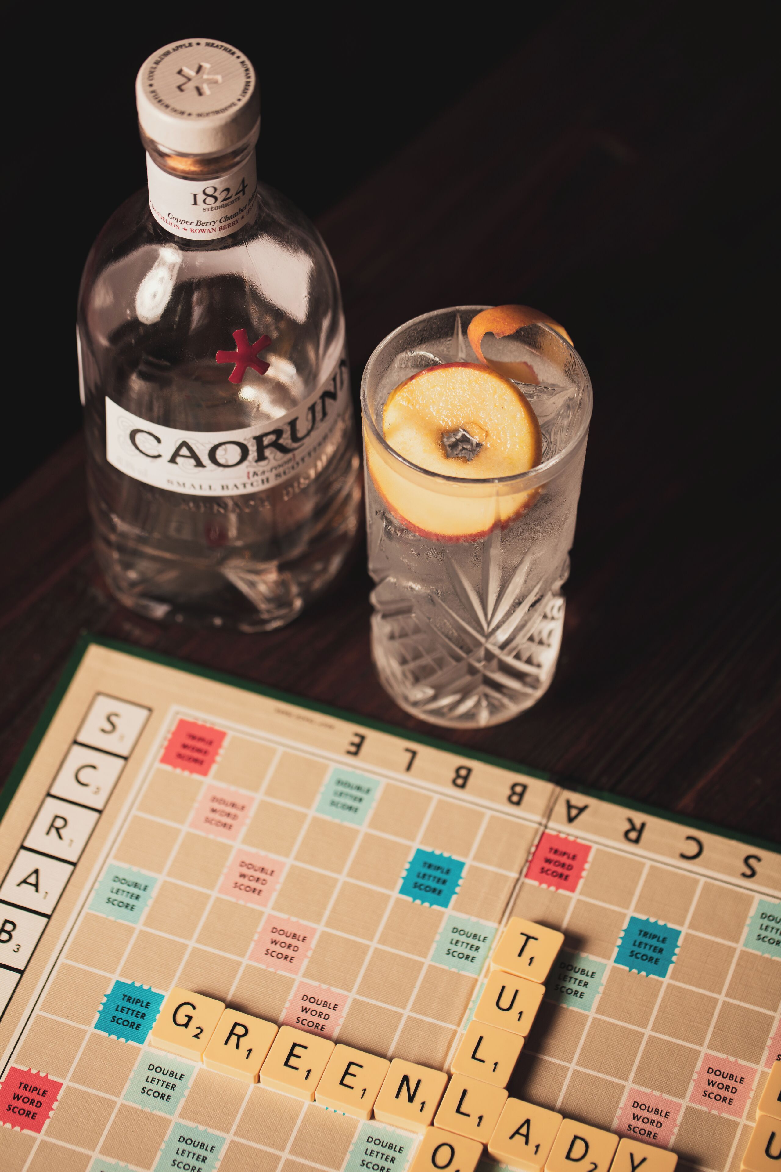 A bottle of Caorun and a glass with a slice of orange on a table with a Scrabble game.