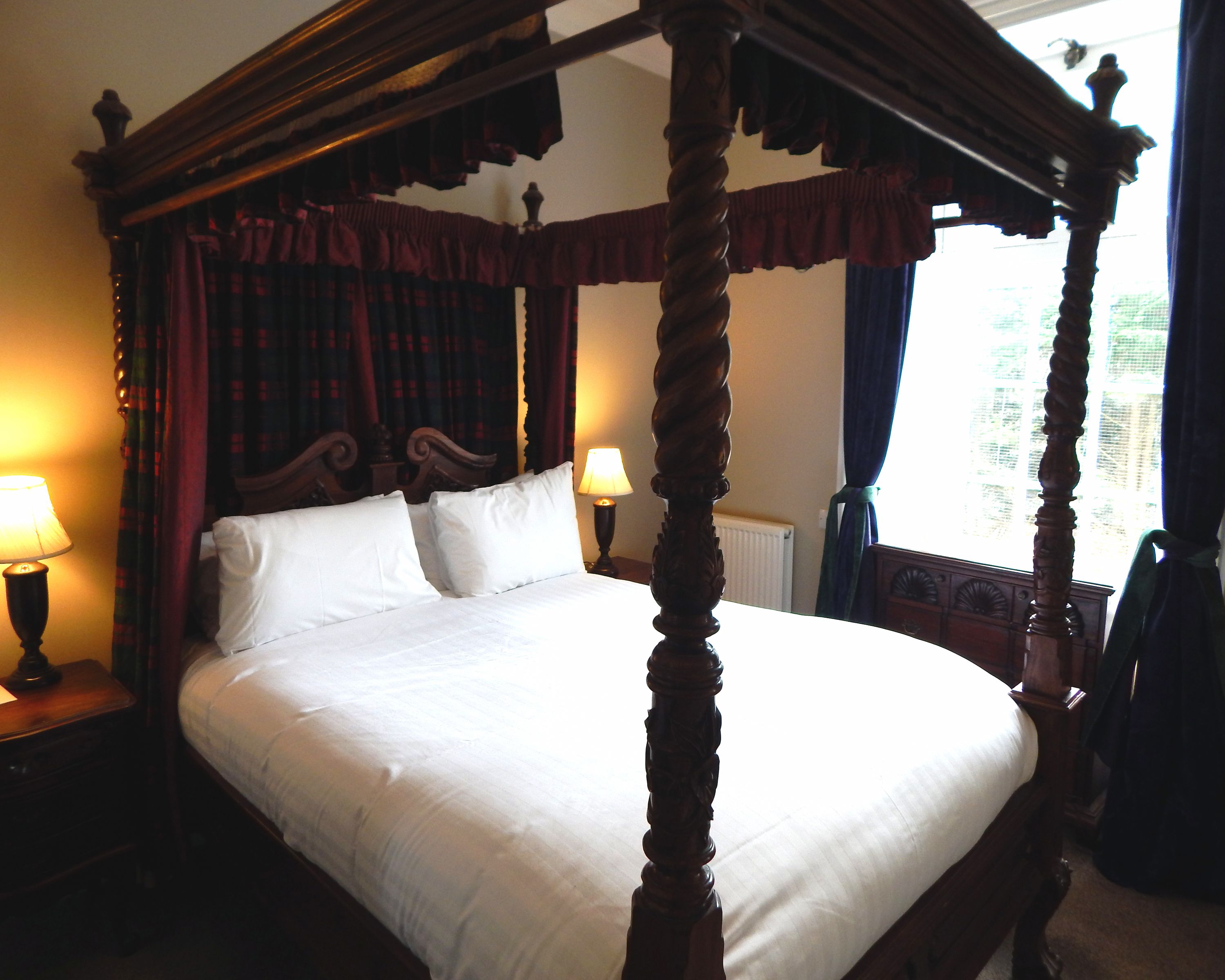 A four-poster bed with white linens, a wooden headboard, and a nightstand with a lamp.