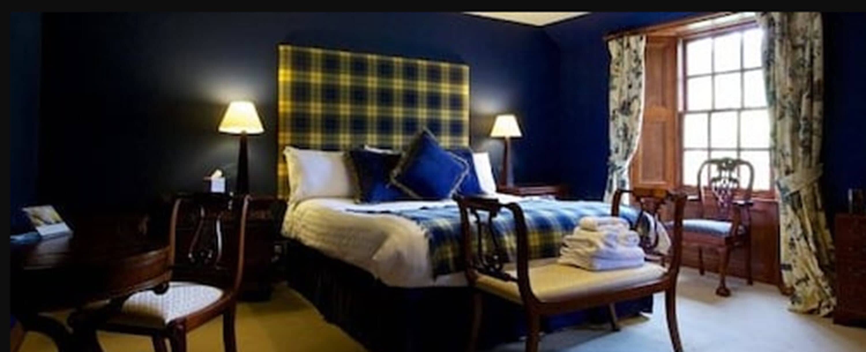 Una habitación de hotel con una cama, dos lámparas de noche, una silla, un escritorio y un ventanal con cortinas.