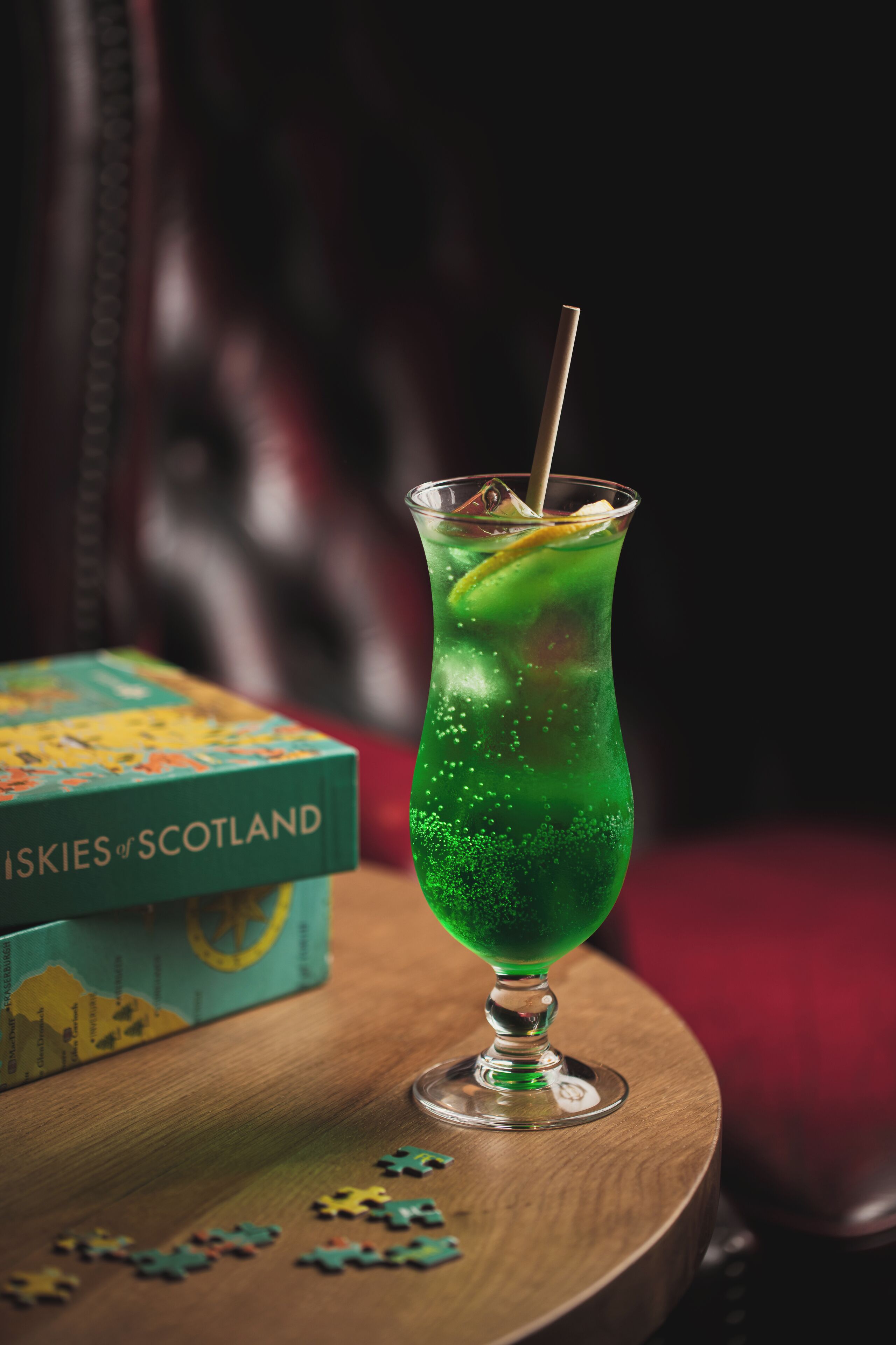 A green cocktail in a glass with a straw, a puzzle box labeled "Scotland," and puzzle pieces on a wooden table.