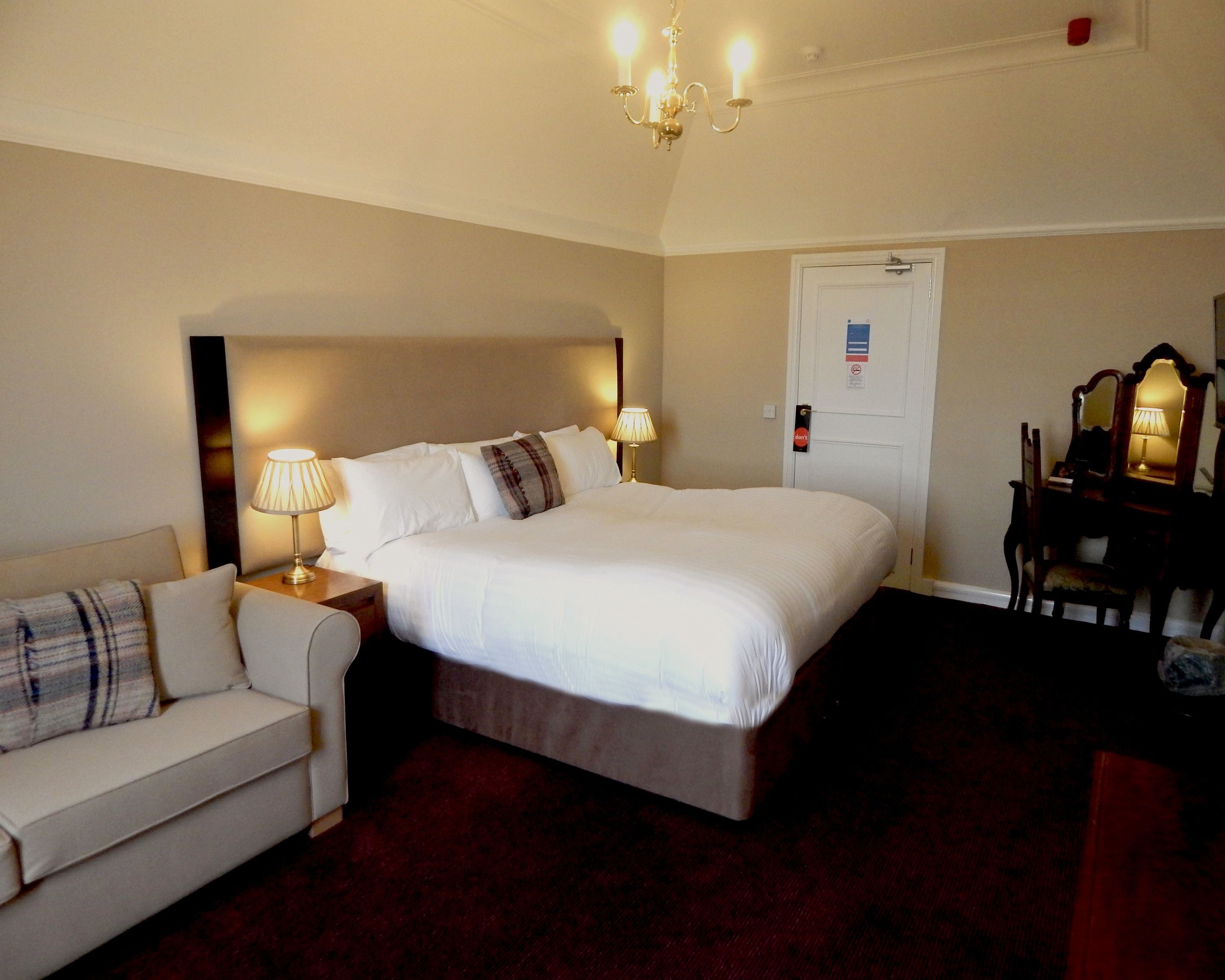 Habitación de hotel con una cama grande, una mesita de noche con lámpara, una silla, un escritorio con espejo y una puerta con letrero.