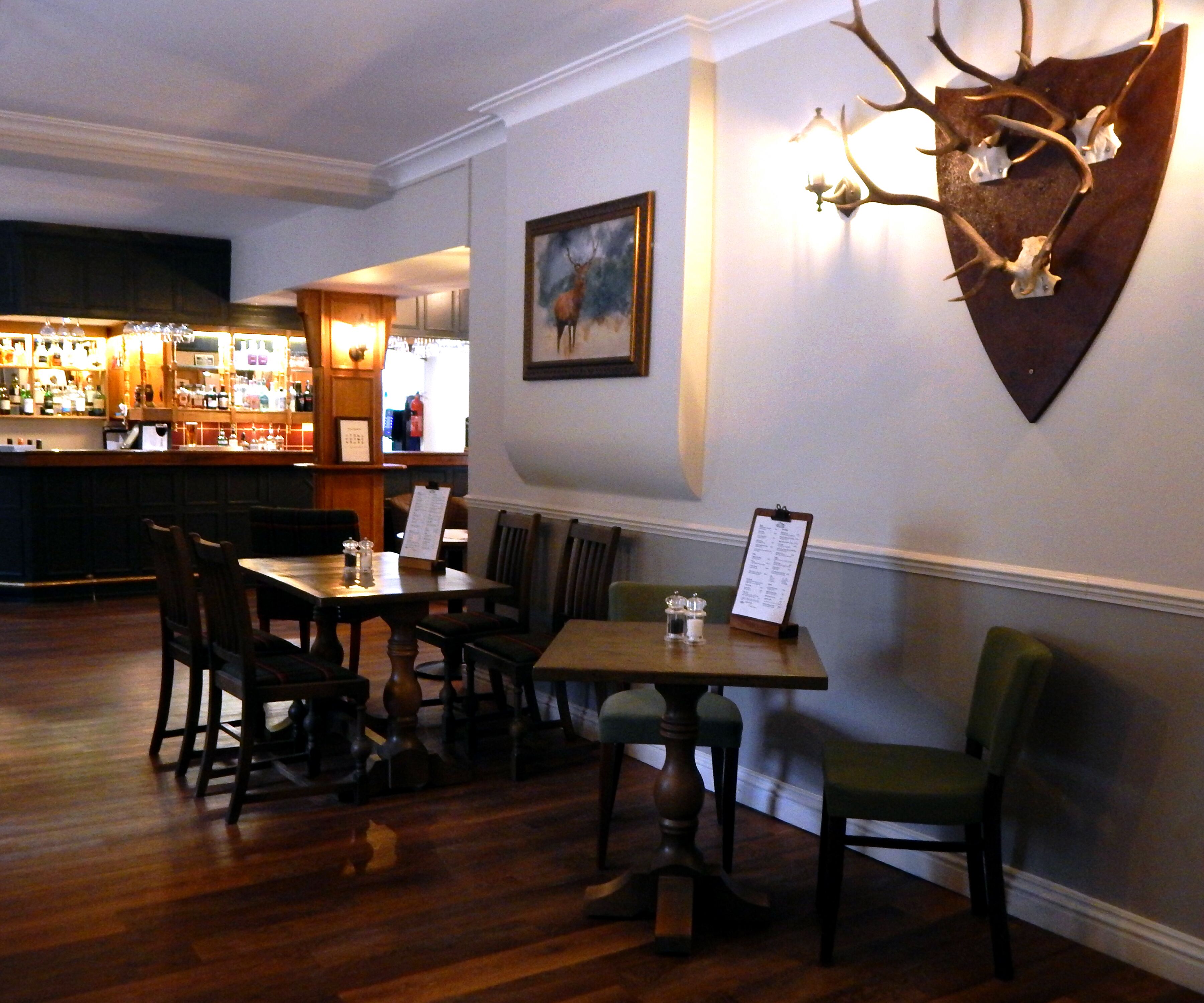 A restaurant interior with wooden tables and chairs, a bar area in the background, and a mounted deer head on the wall.