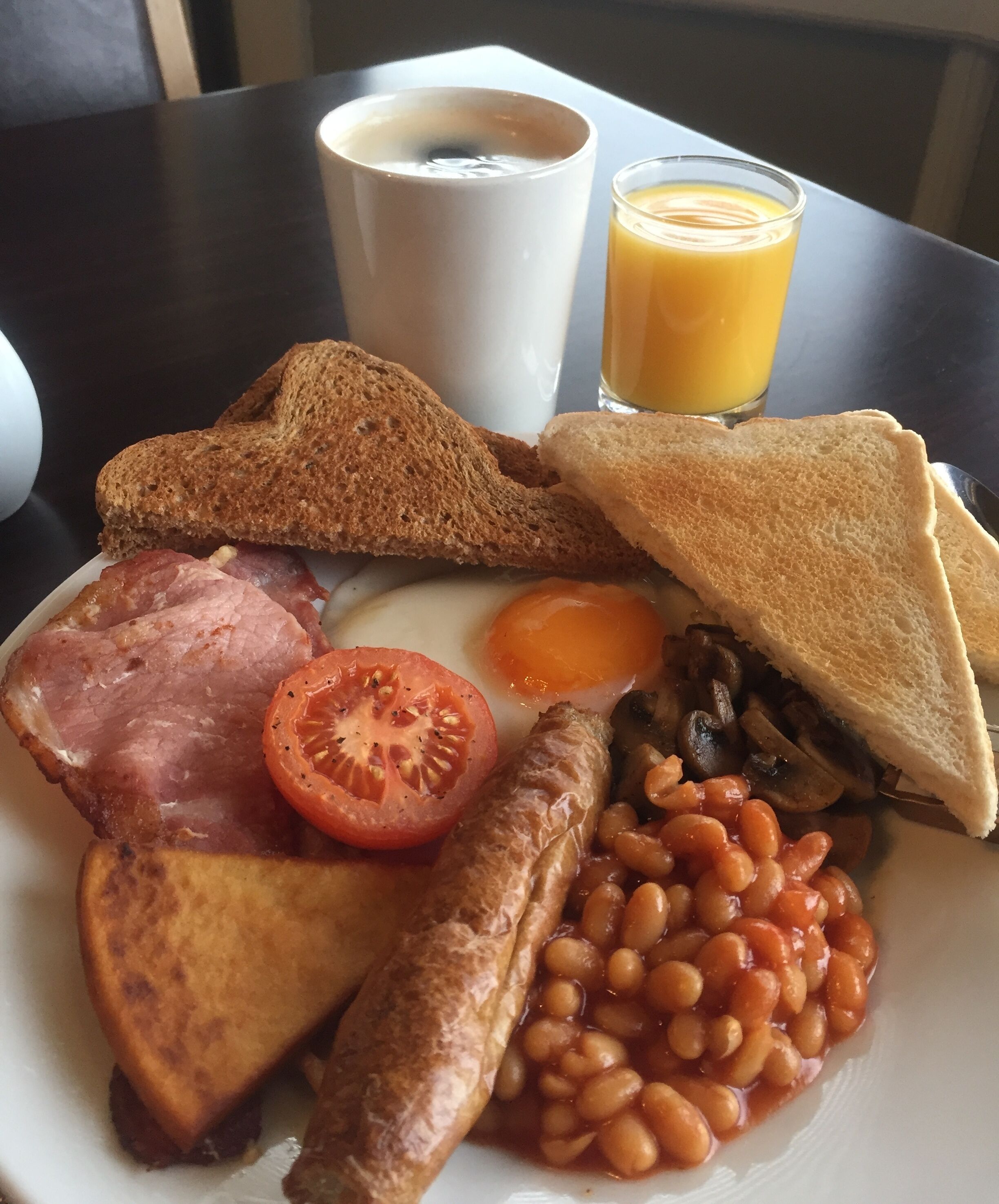 Une assiette de nourriture pour le petit-déjeuner comprenant des toasts, des œufs, du bacon, des tomates, des haricots et un verre de jus d’orange.