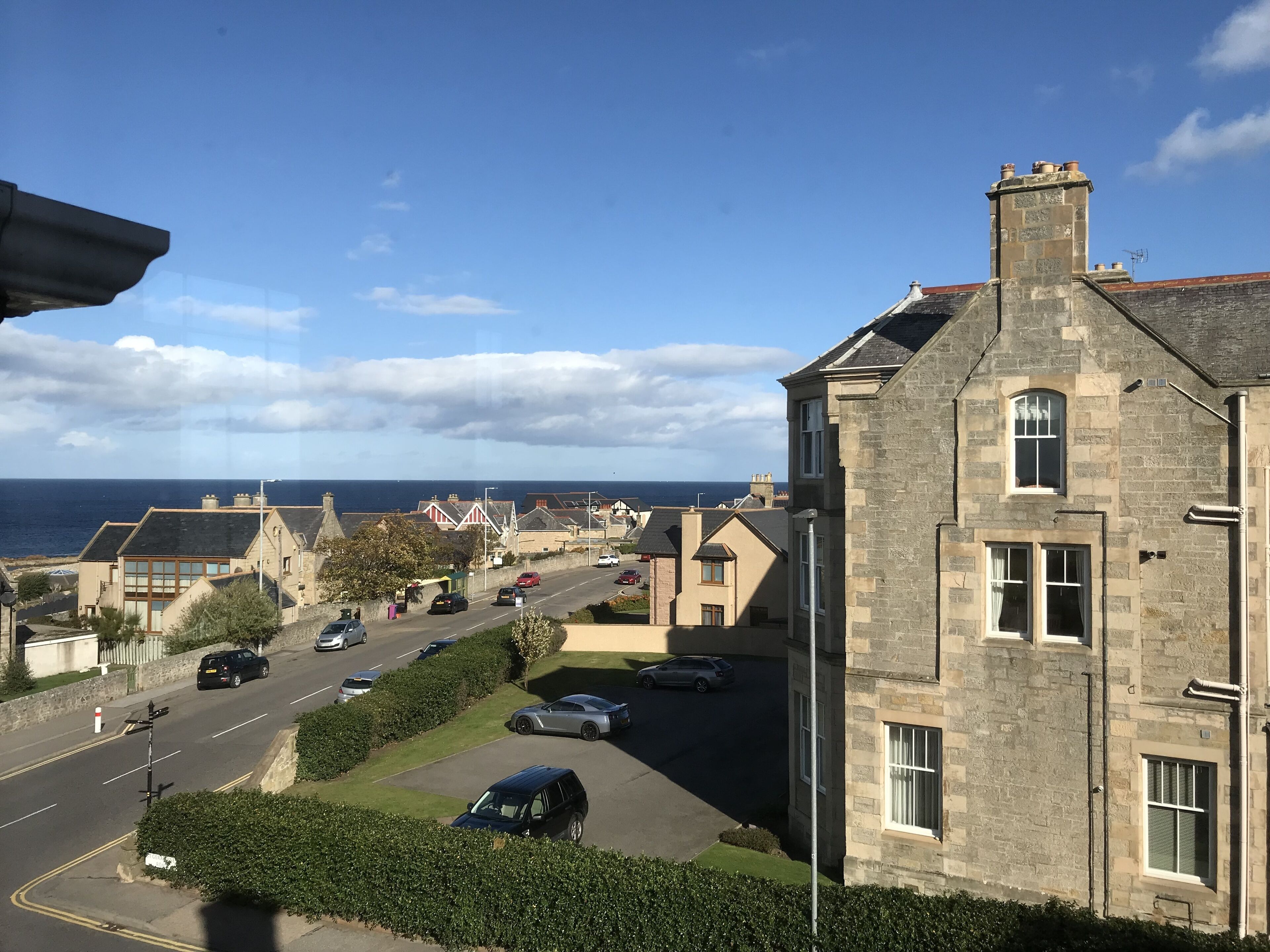 A coastal residential area with stone buildings, a road with parked cars, and a view of the sea.
