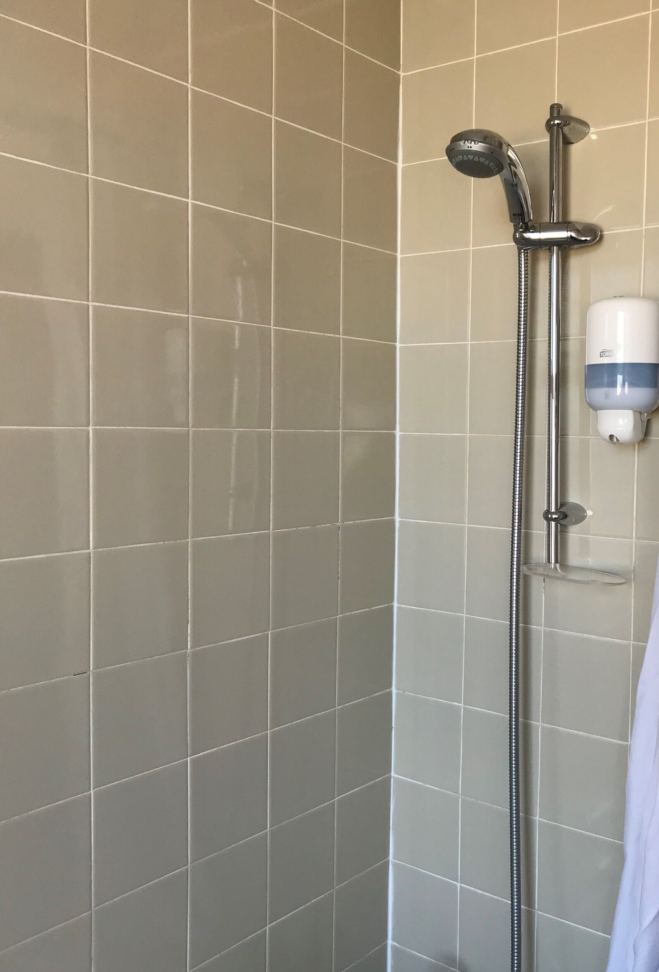 A corner of a bathroom with beige tiled walls and a shower area with a handheld showerhead and a wall-mounted soap dispenser.