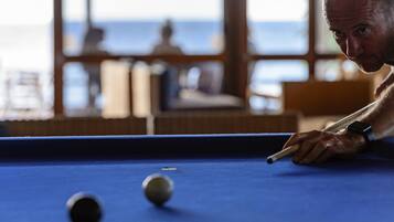 Une table de billard avec une personne penchée au-dessus, une nappe bleue et, à l’arrière-plan, une vue sur la mer à travers de grandes fenêtres.