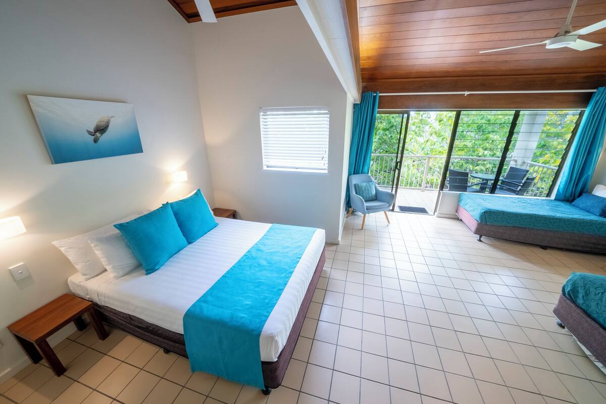 A bedroom with a bed, a nightstand, a window with blinds, a balcony with a chair, and a view of greenery.