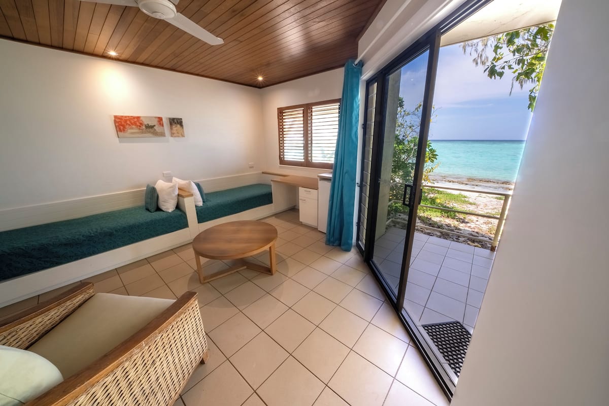 A room with a bed, a small table, a chair, a bench, and a view of the beach through a sliding glass door.