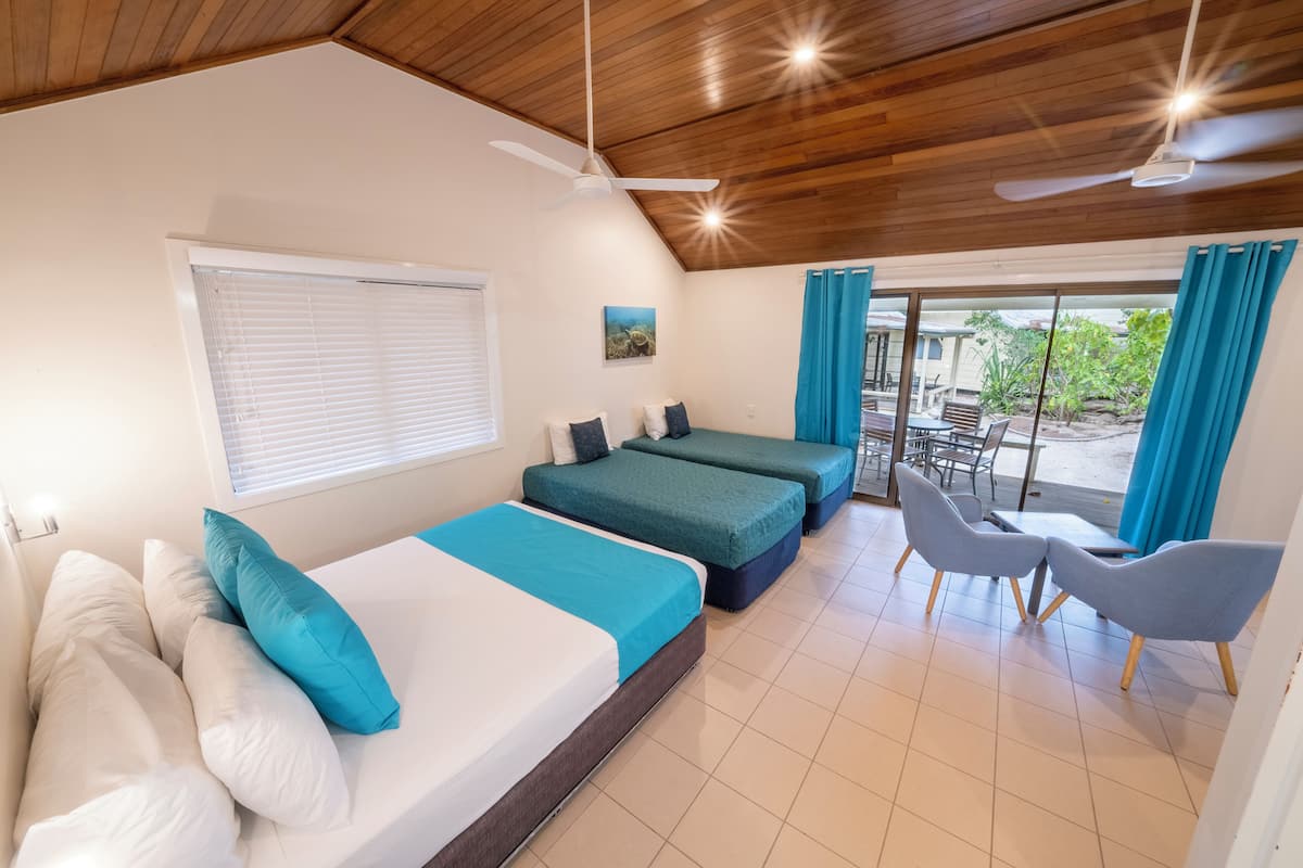 A room with two beds, a ceiling fan, a window with blinds, a dining area with chairs, and a view of the outdoors.