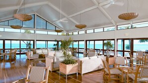 A spacious indoor area with multiple seating arrangements, large windows offering ocean views, and hanging wicker lamps.