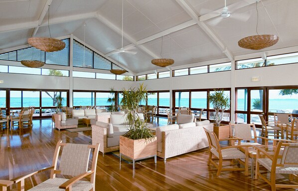 A spacious indoor area with multiple seating arrangements, large windows offering ocean views, and hanging wicker lamps.