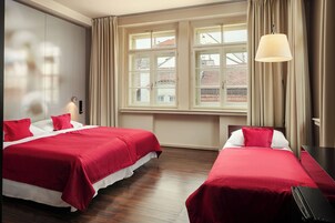 Quarto de hotel com duas camas, uma janela grande com cortinas, uma escrivaninha e uma luminária.