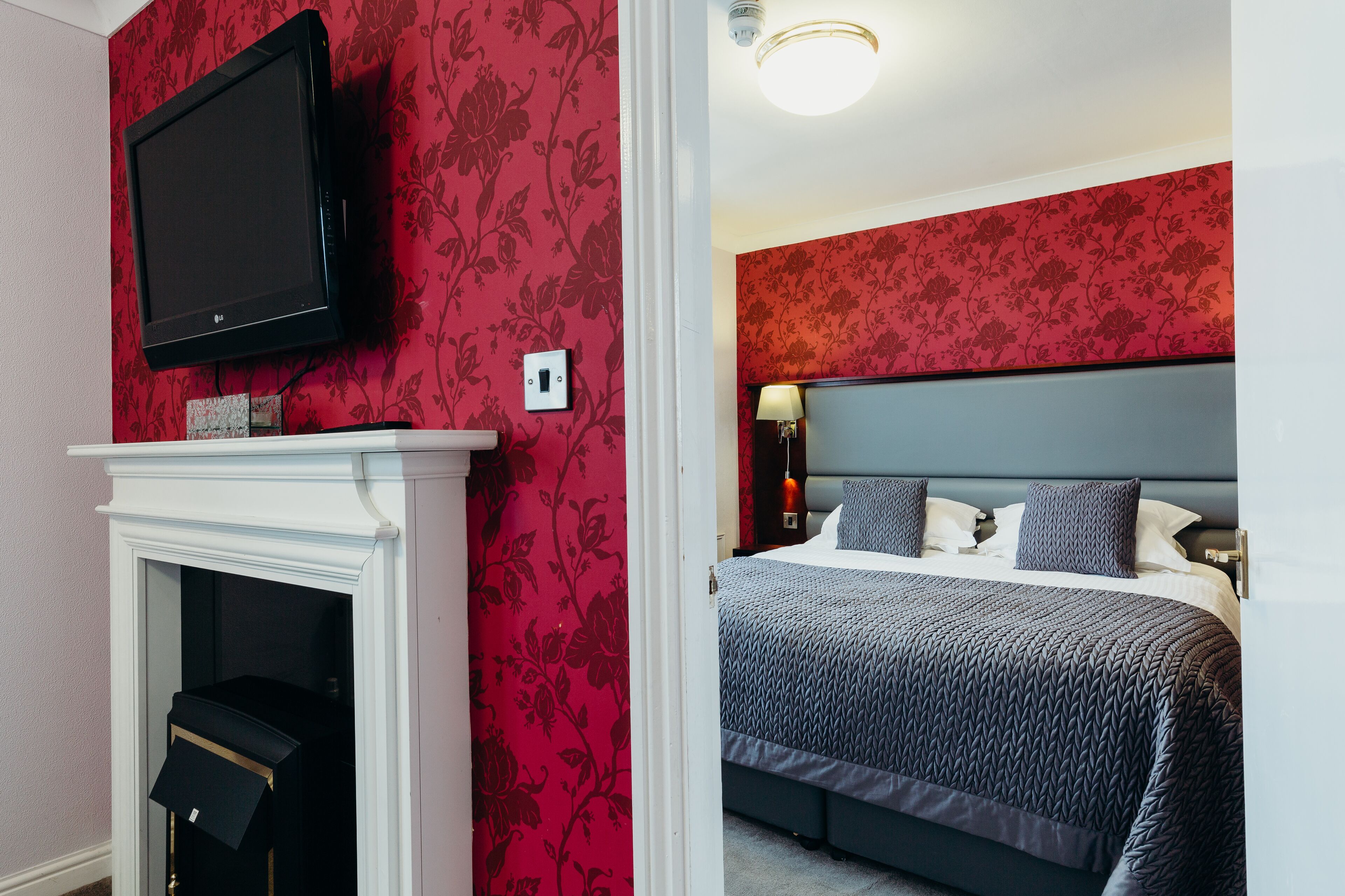 Quarto com uma cama grande, uma televisão montada acima de uma lareira e um papel de parede floral vermelho.