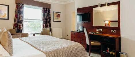 A hotel room with a large bed, a desk with a TV, a chair, and a bookshelf.