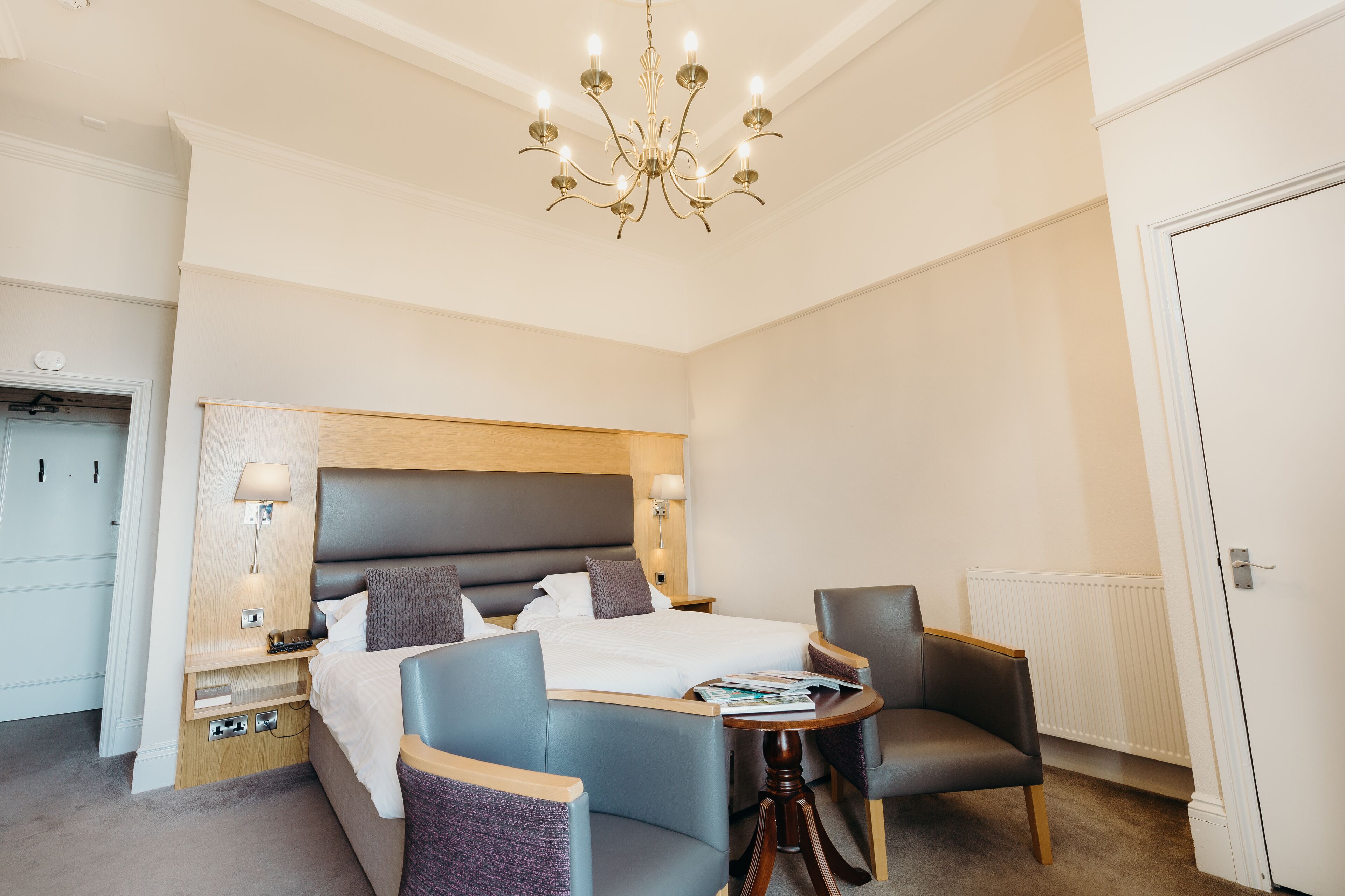 Une chambre d’hôtel avec un grand lit, deux fauteuils, une petite table ronde, un lustre et une table de chevet avec une lampe.