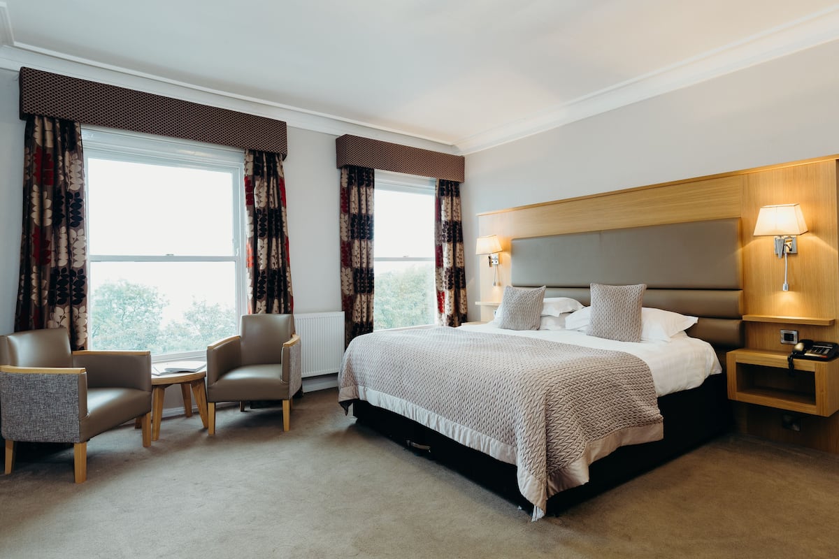 Habitación de hotel con una cama grande, dos sillones, una mesita y una mesita de noche con lámpara.
