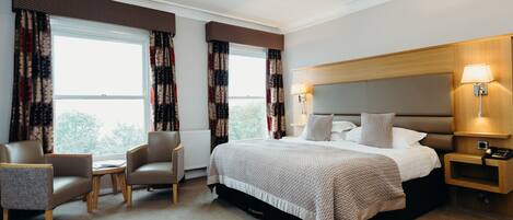 A hotel room with a large bed, two armchairs, a small table, and a nightstand with a lamp.