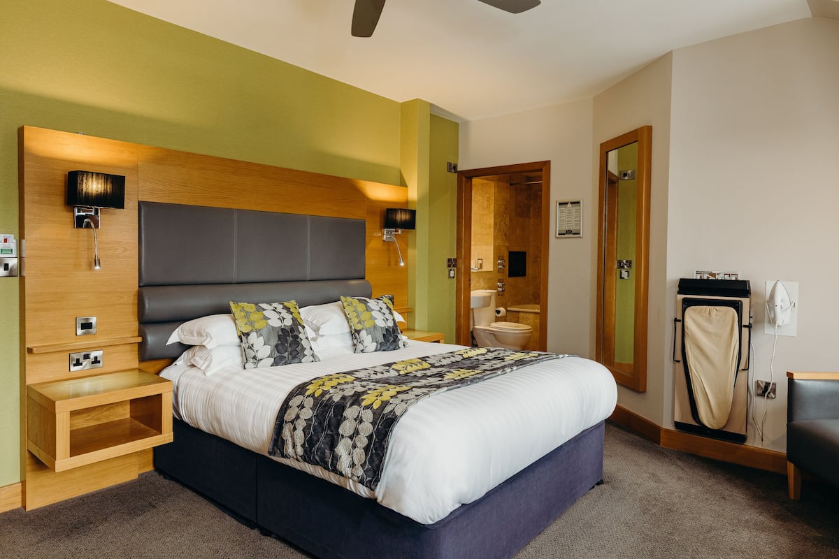 Habitación de hotel moderna con una cama grande, cabecera de madera, mesitas de noche, un espejo y una pequeña zona de estar.