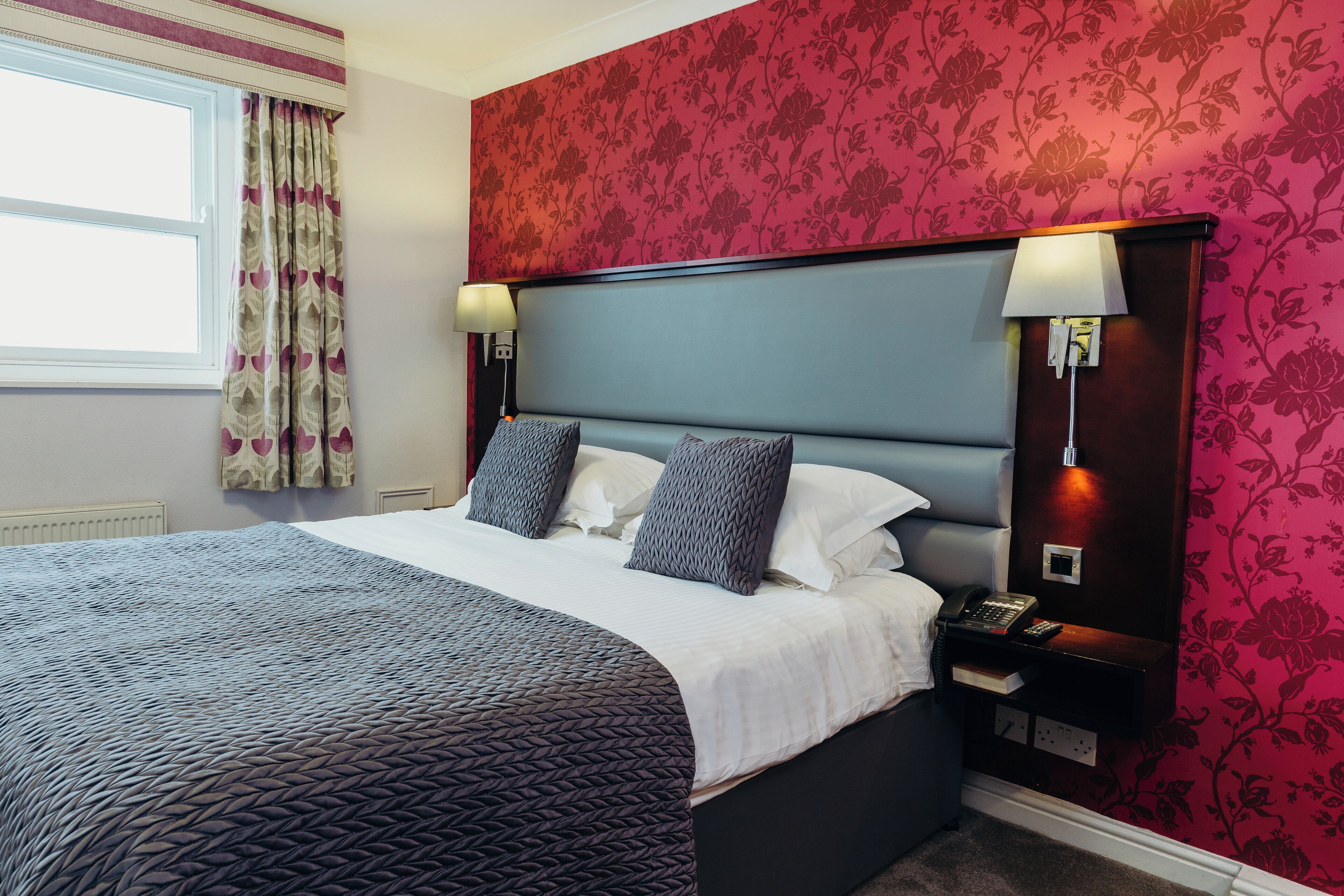 A neatly made bed with a patterned bedspread, a headboard with two wall-mounted lamps, and a telephone on a bedside table.
