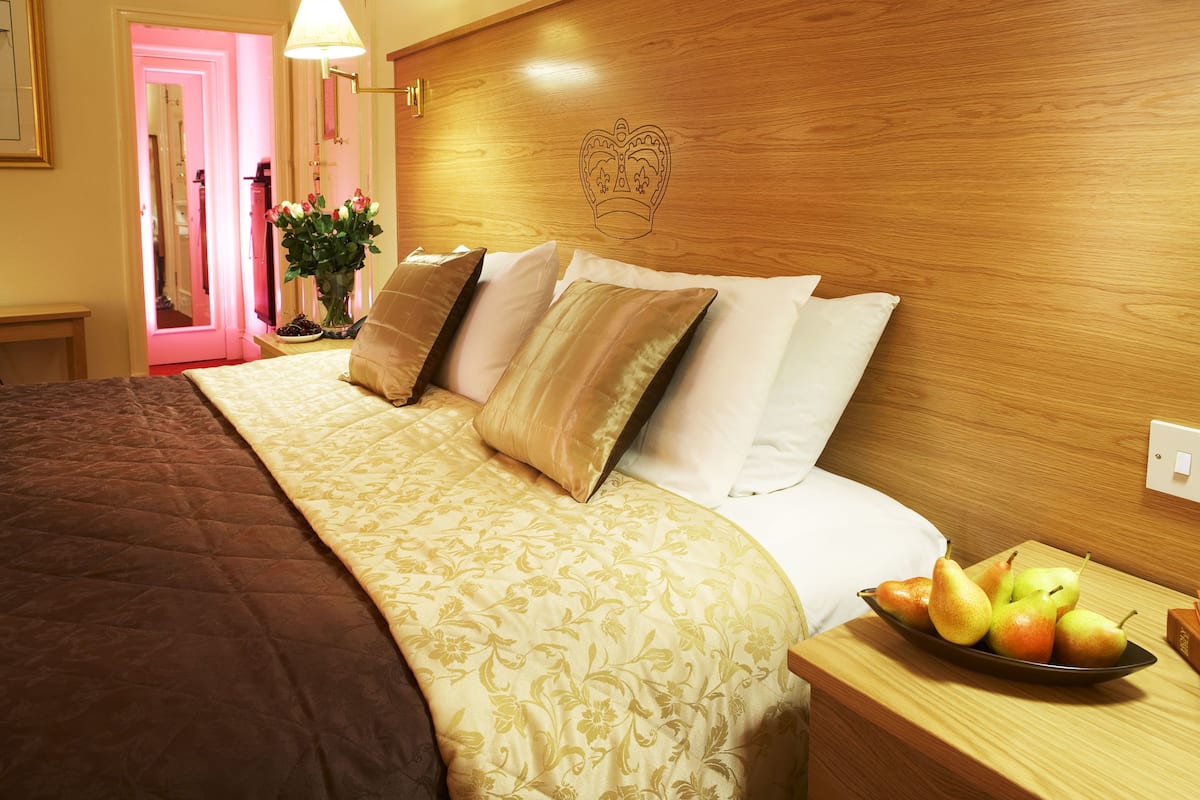 Una cama bien hecha con una colcha estampada con motivos florales, una mesita de noche con un frutero y un cabecero de madera con un diseño de corona.