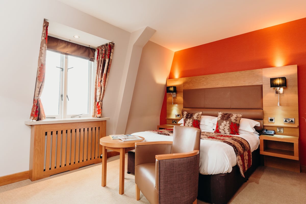 Habitación de hotel con cama, una silla, una mesa, una mesita de noche y una ventana con cortinas.