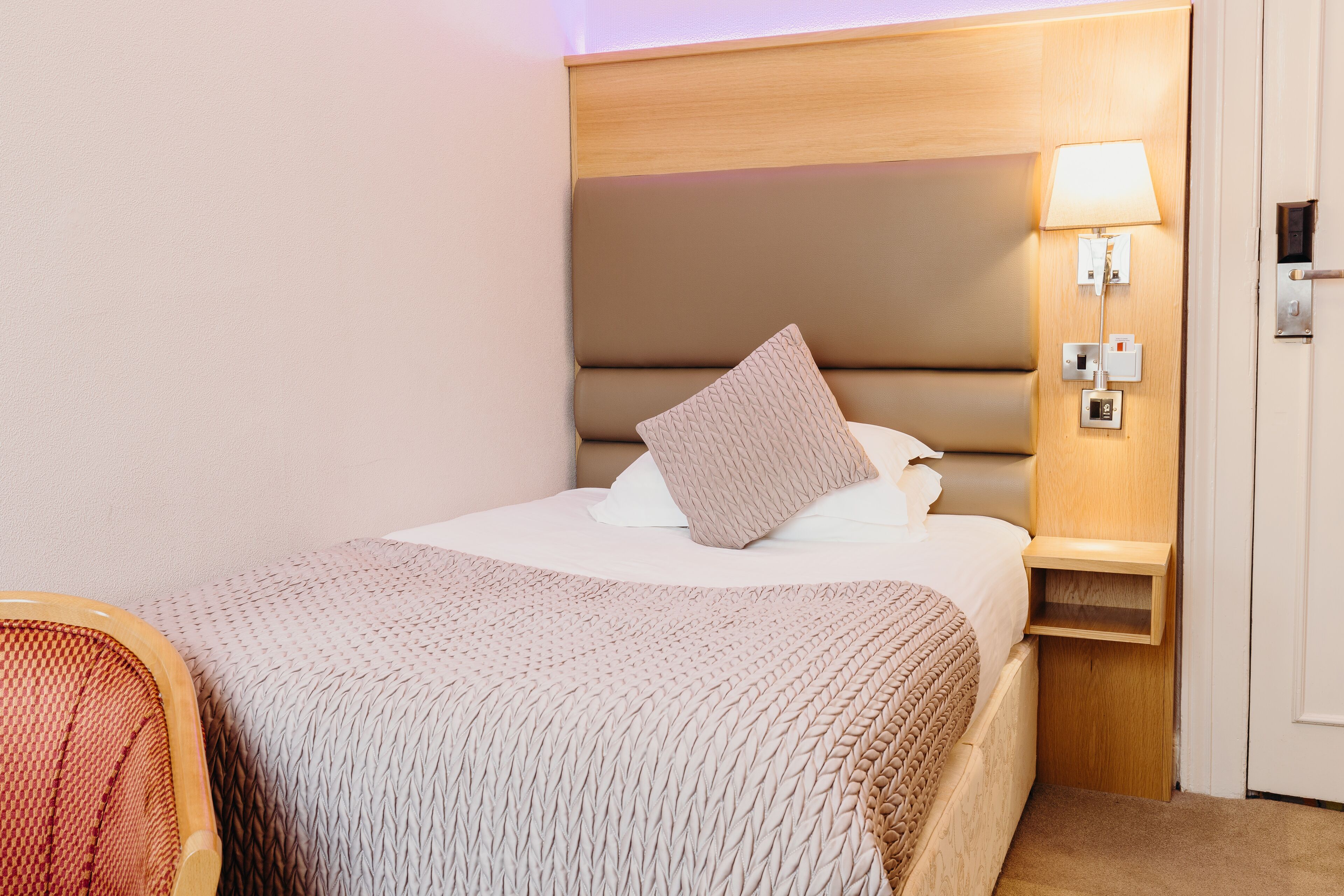 A neatly made bed with a headboard, a bedside table with a lamp, and a chair with a patterned cushion.