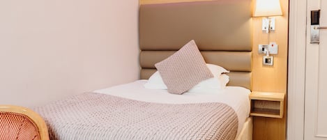 A neatly made bed with a headboard, a bedside table with a lamp, and a chair with a patterned cushion.