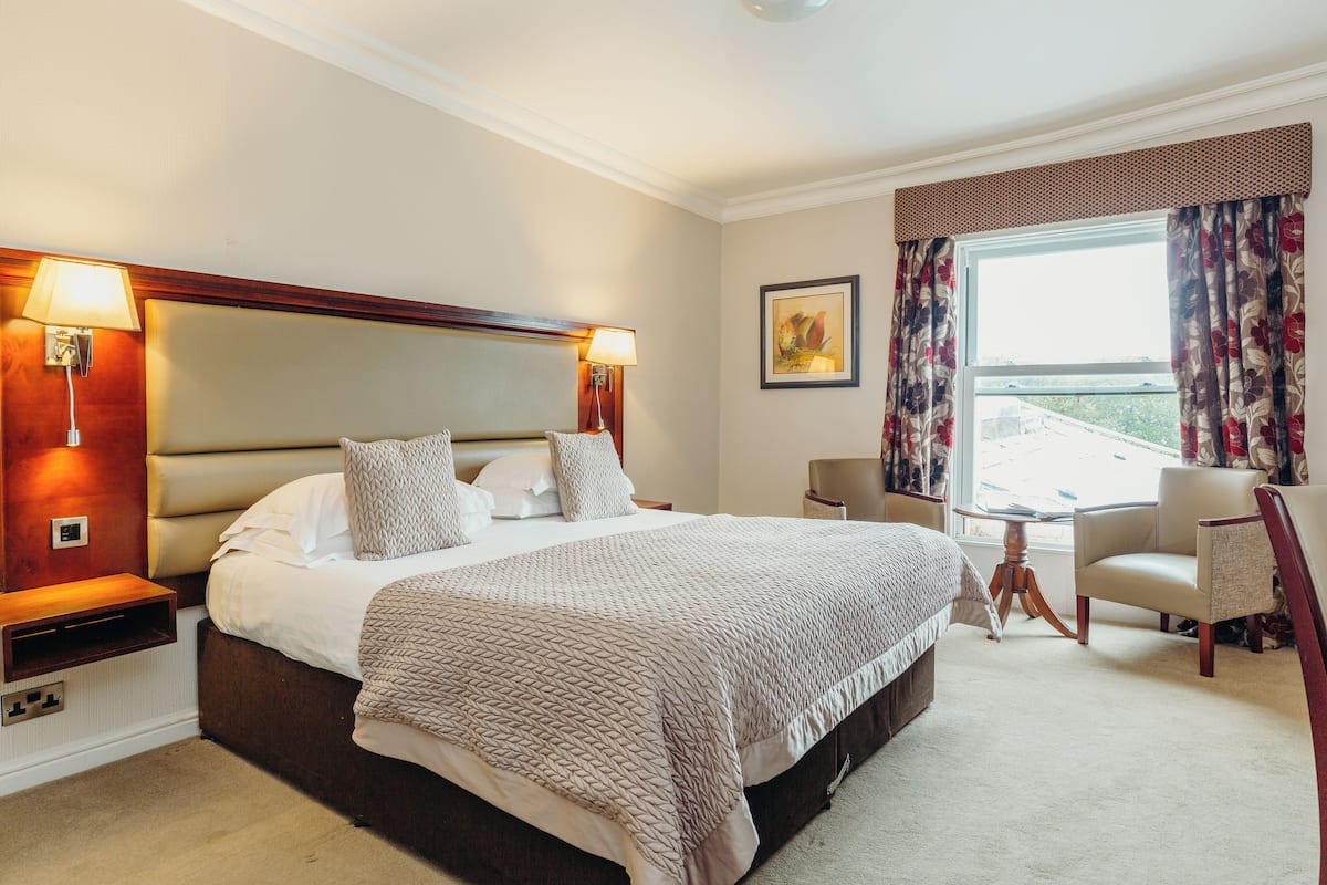 Habitación de hotel con una cama grande, dos lámparas de noche, una silla, una mesita y una ventana con cortinas.