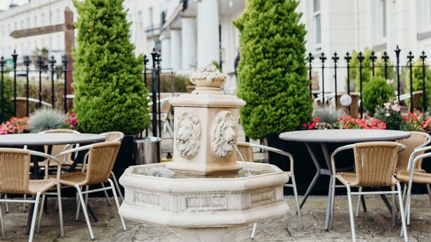 A stone fountain with two lion sculptures, surrounded by wicker chairs and a table, set against a backdrop of a white building with arched windows and a black wrought iron fence.