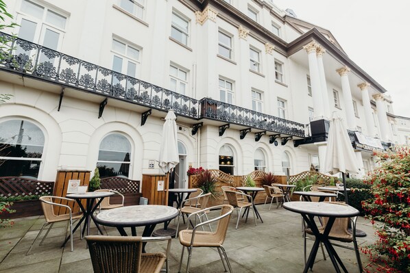 An outdoor cafe area with tables and chairs, a wooden fence, and a building with columns and arched windows.