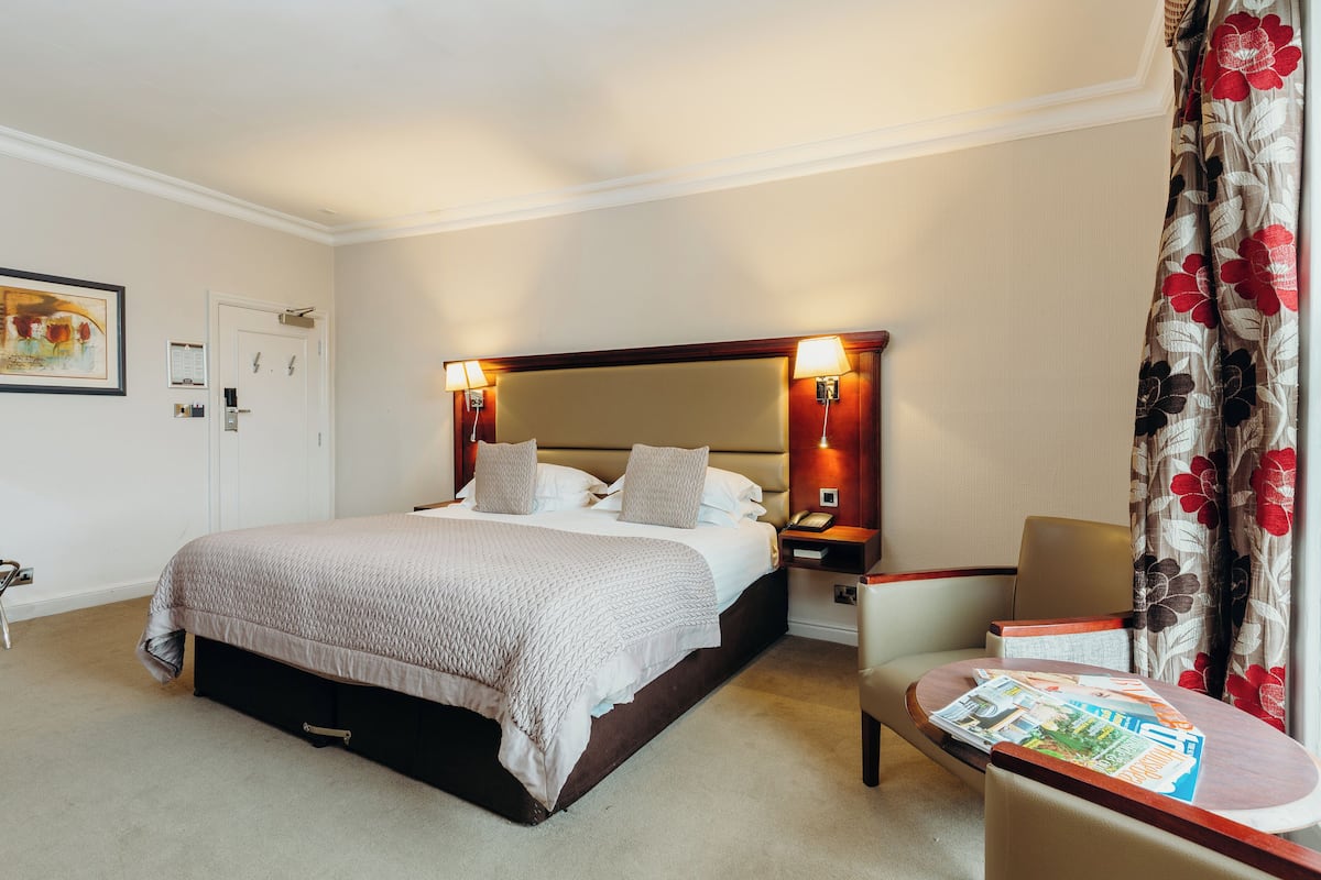 Una habitación de hotel con una cama grande, lámparas de noche, una silla y una mesa con revistas.