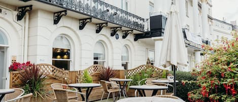 An outdoor cafe area with wicker tables and chairs, a wooden fence, and a balcony with ornate metal railings.