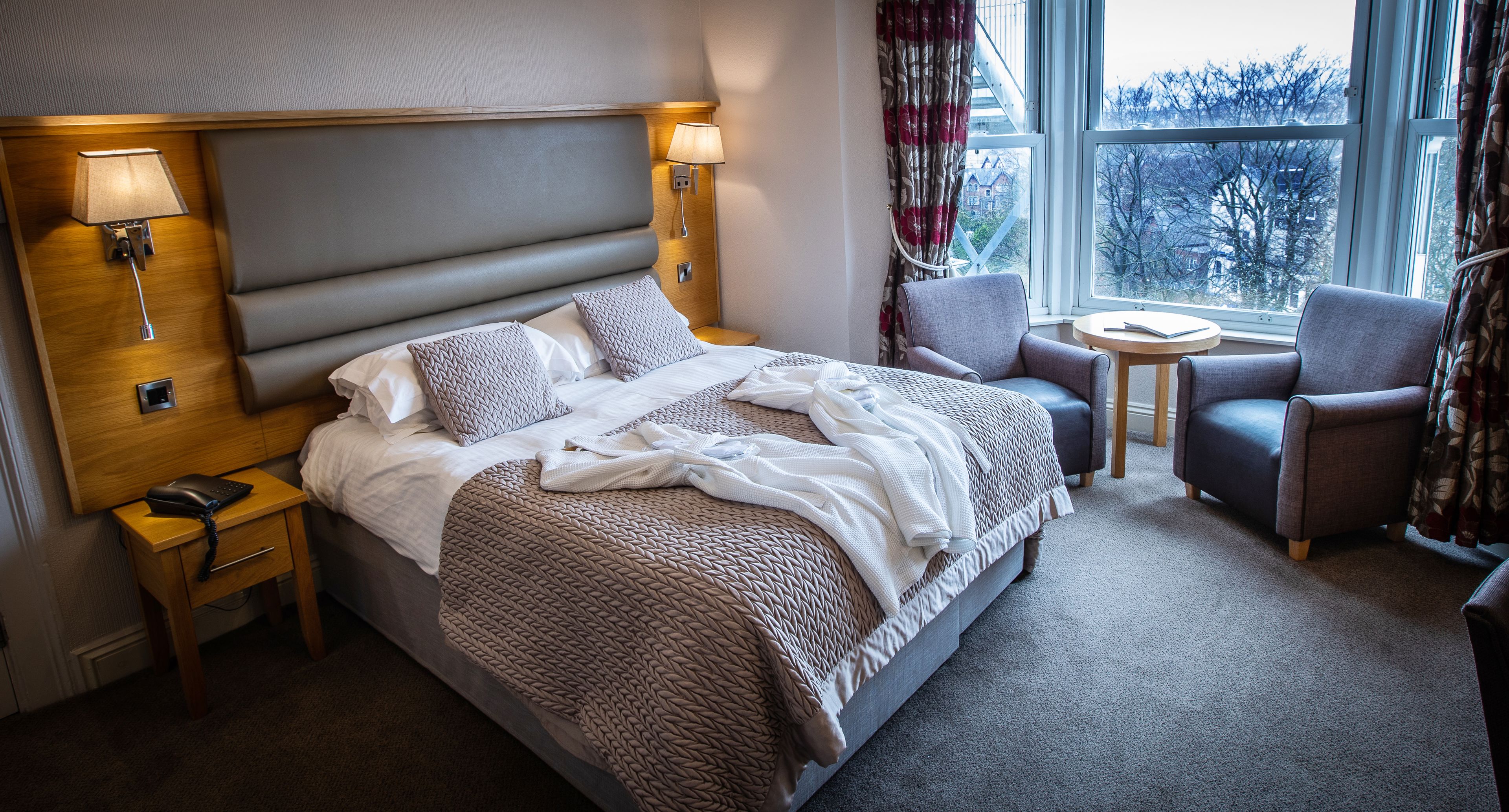 Ett hotellrum med en stor säng, två sänglampor, ett litet bord, en stol och utsikt över omgivningarna genom ett fönster.