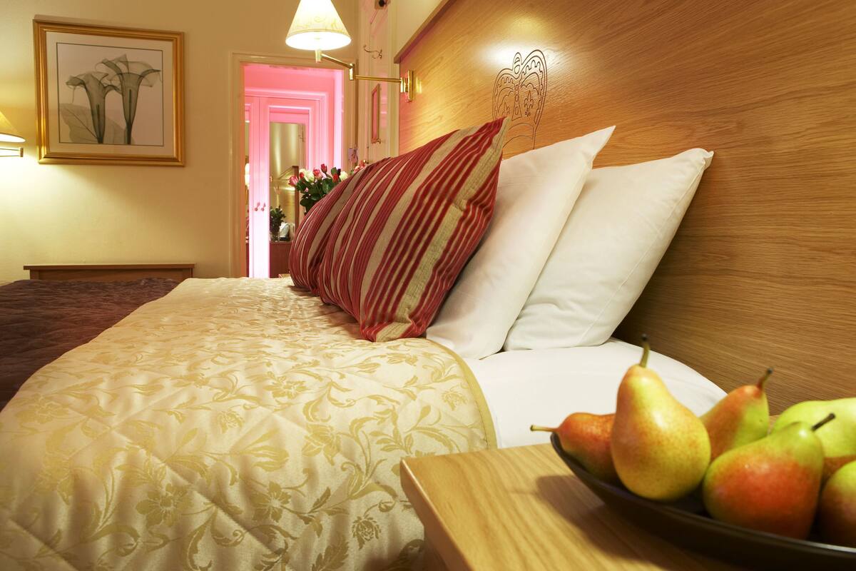 Una habitación de hotel con una cama, cabecero con diseño de escudo, mesita de noche con frutero y un cuadro en la pared.