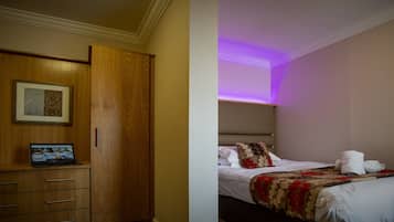 Habitación de hotel con un armario de madera, un ordenador portátil sobre la mesa, una cama con colcha estampada y una luz morada en el techo.