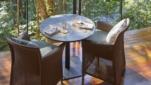 A round table with two wicker chairs and a glass on it, set on a balcony with a view of a lush garden and a stream. - Silky Oaks Lodge, Beckons (Finlayvale)