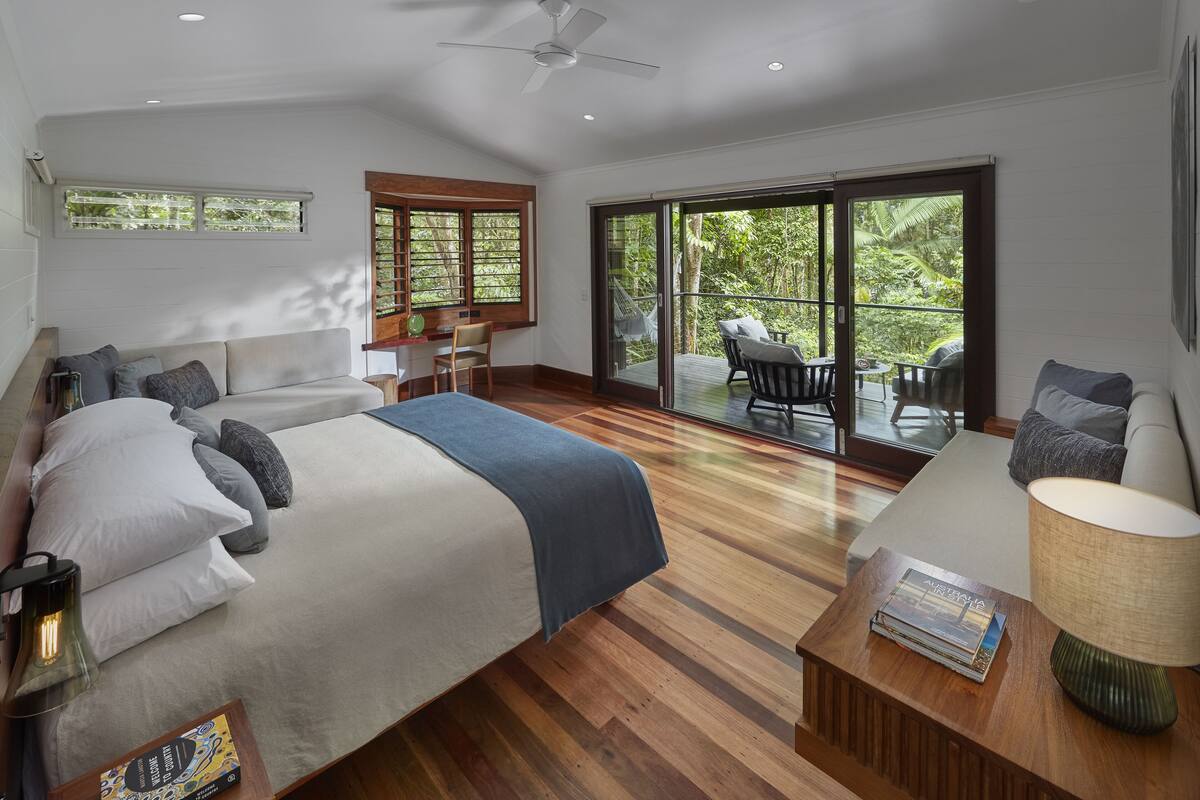 A spacious bedroom with a large bed, a sitting area with a table and chairs, and a balcony with a view of the greenery.