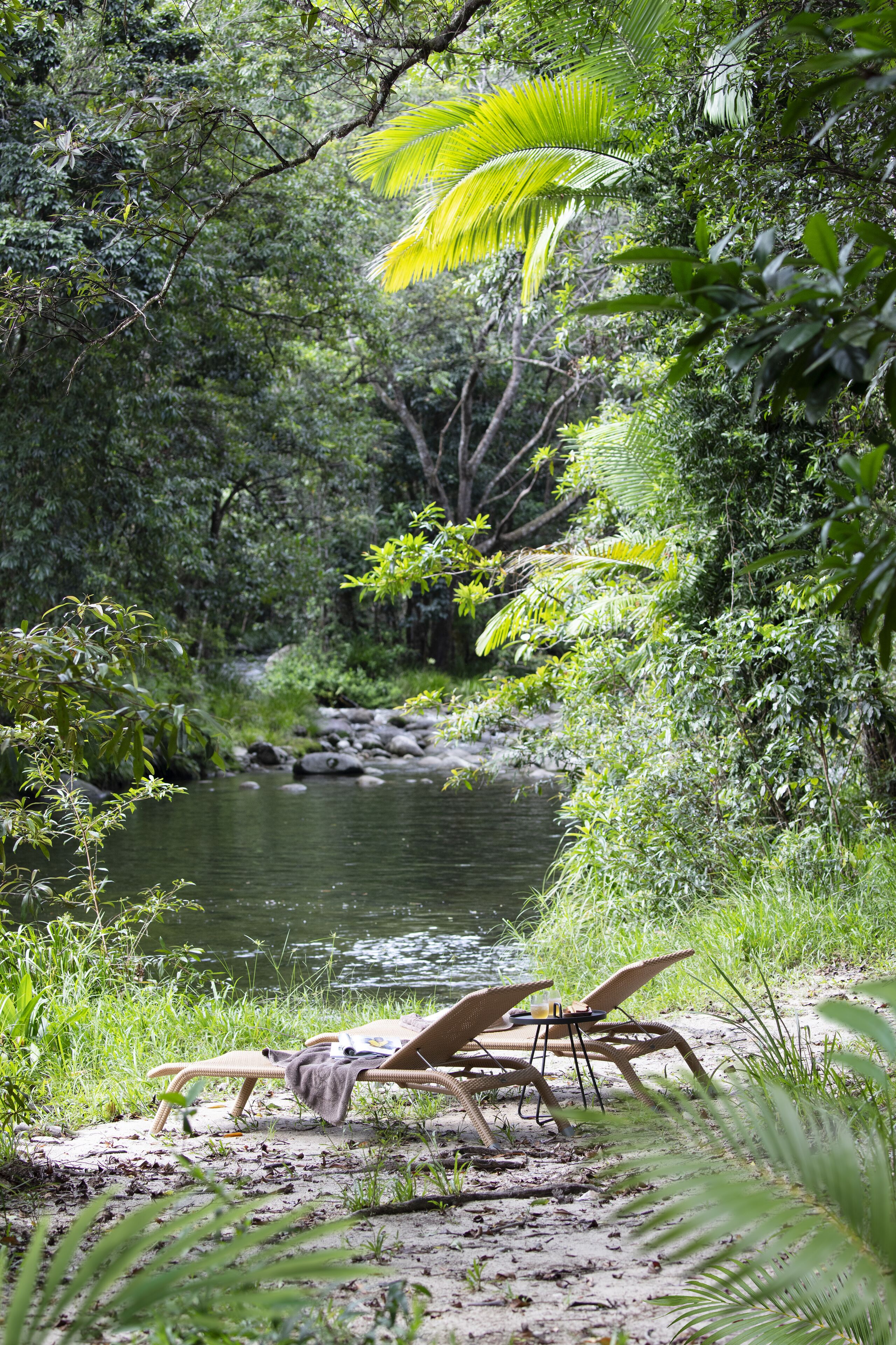 A serene riverside setting with a wicker lounge chair and a table, surrounded by lush greenery and a calm river.