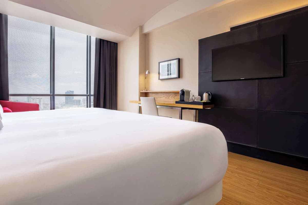 A hotel room with a large bed, a desk with a chair, a television mounted on the wall, and a window offering a city view.