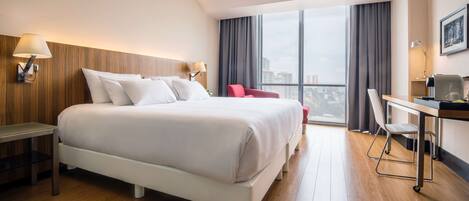 A hotel room with a large bed, a desk, a chair, and a view of the city through large windows.