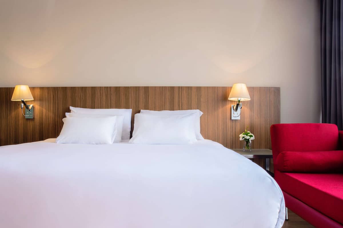 A hotel room with a large bed, two bedside lamps, a red armchair, and a small table with a vase of flowers.