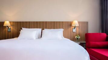 Una habitación de hotel con una cama grande, dos lámparas de noche, un sillón rojo y una mesita con un jarrón de flores.