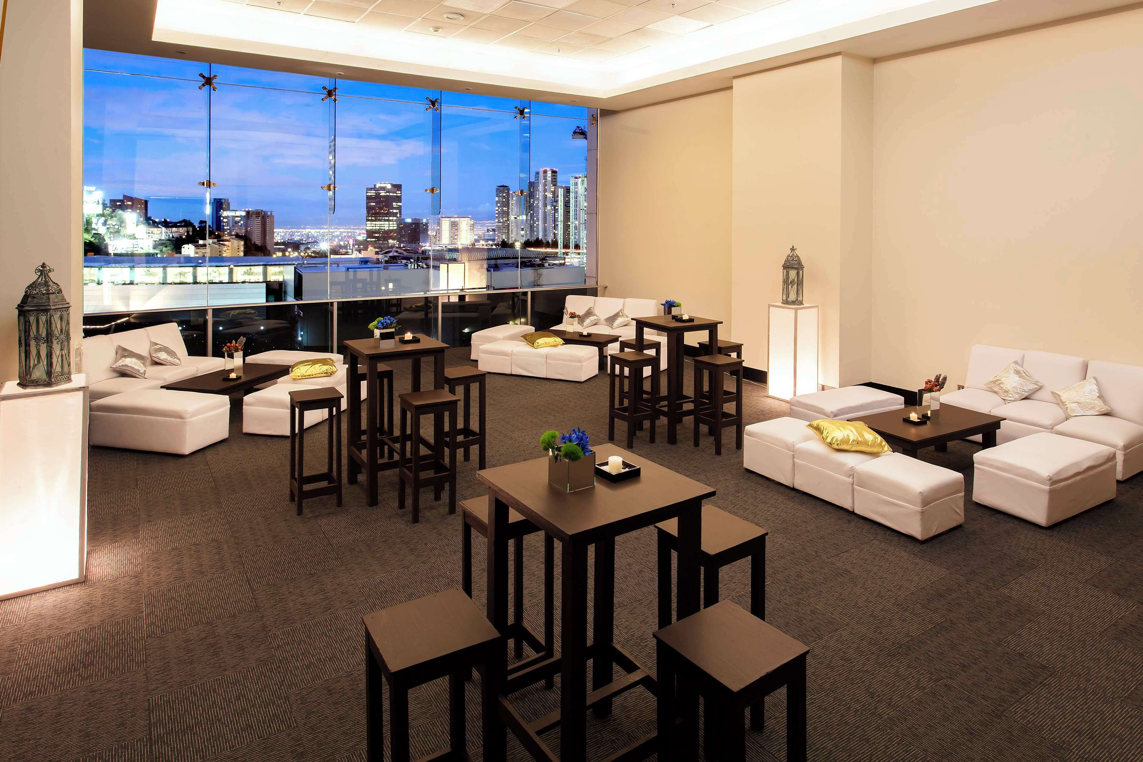 A modern lounge with white sofas, dark wooden tables, and a city view through large windows.