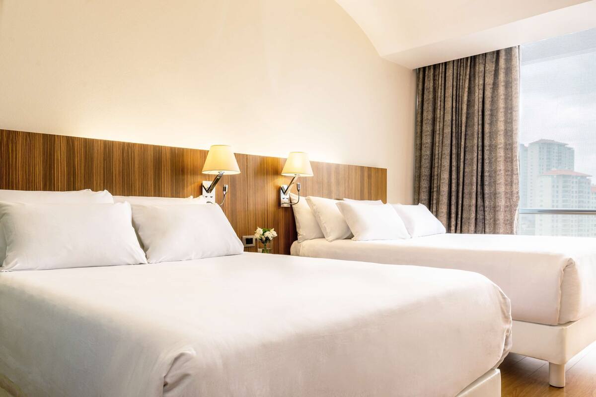 A hotel room with two beds, a wooden headboard, two wall-mounted lamps, and a large window with curtains.