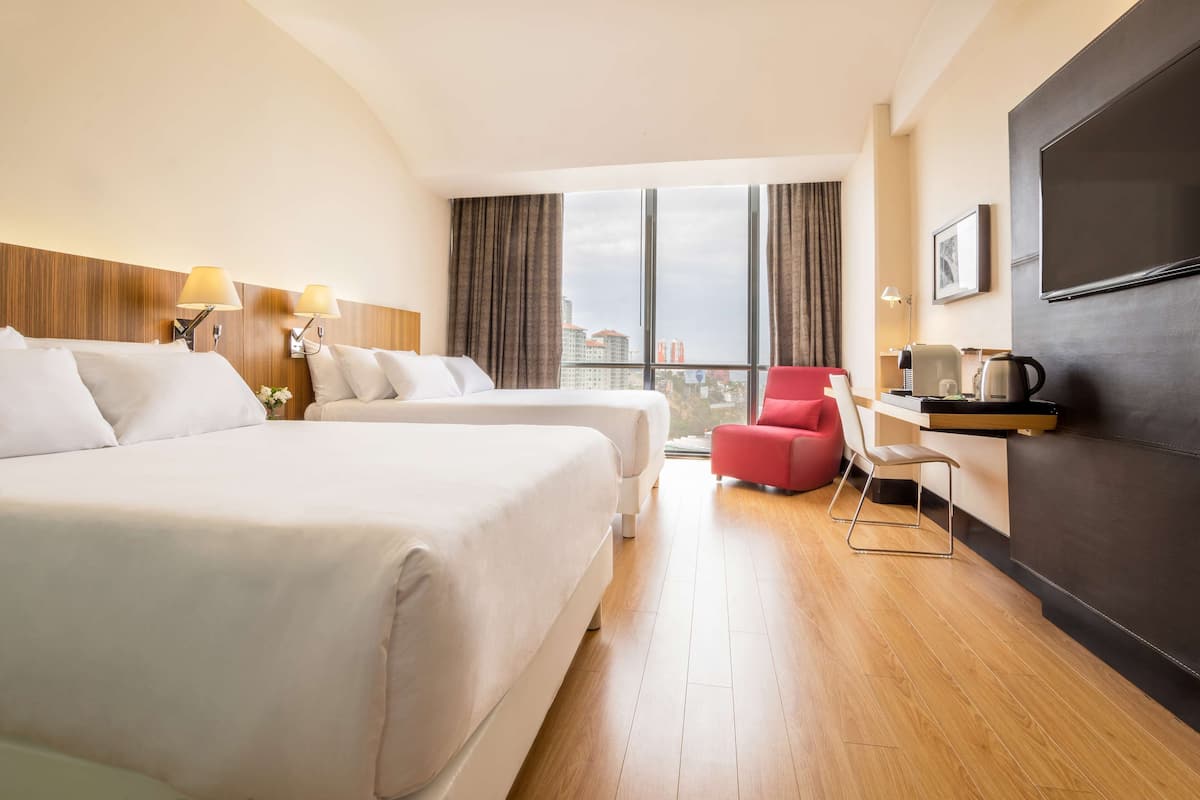 A hotel room with two beds, a desk, a red chair, and a large window offering a city view.
